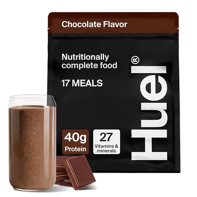A bag of Huel Chocolate Flavor protein powder shown next to a glass of chocolate-colored shake and chocolate pieces on a white background.