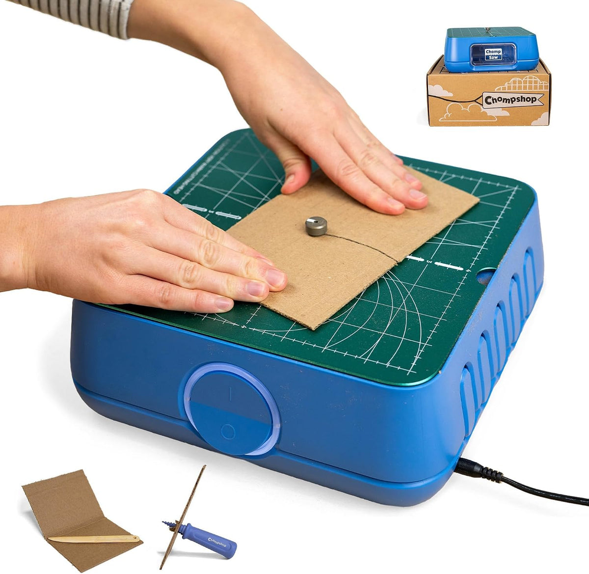 Hands guiding cardboard on a blue Chompshop ChompSaw cutter with a green gridded work surface and power cord